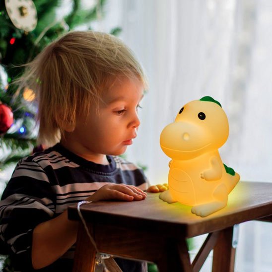 Veilleuse bébé dinosaure vert Dino Câlin allumée sur une table de chevet